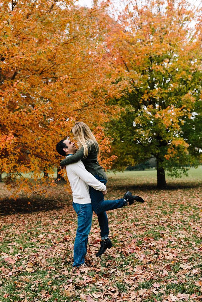 pose idea for couple, guy picks up girl and she kicks her foot out and looks down at him in front of brightly colored fall trees in the midwest