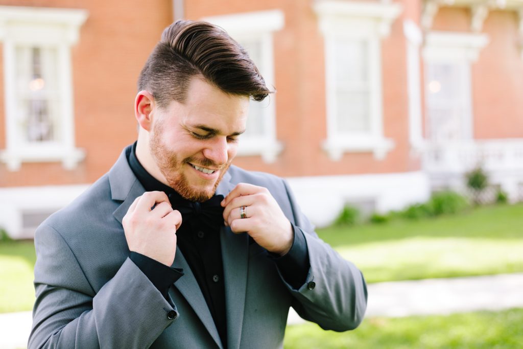 Kansas City groom adjusting his black tie. He wears a grey suit with a black shirt and mixed metal wedding band