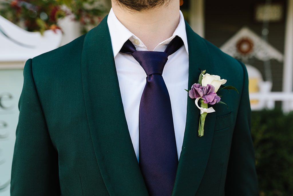 groom wears an emerald green suit jacket with a purple tie from The Black Tux and has a white rose and purple flower boutonniere