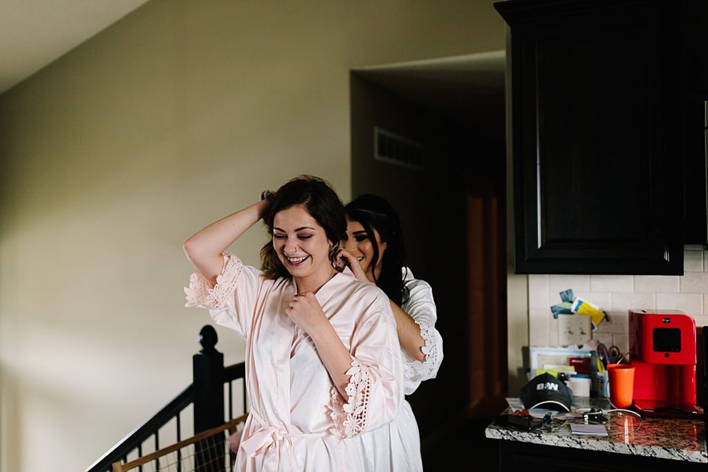 bride helps bridesmaid put on her necklace and they share a candid laugh together