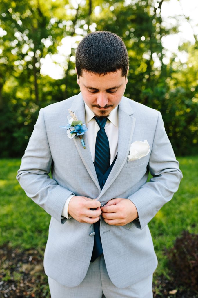 groom buttons his grey suit jacket while wearing a navy tie and handmade boutonniere