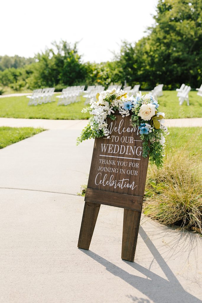 wooden welcome to our wedding sign decorated with white flowers, blue flowers, and greenery for an outdoor wedding ceremony
