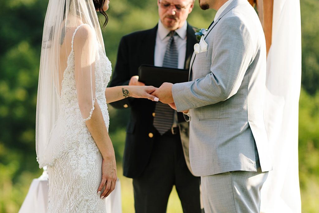 groom places wedding band on bride finger during their outdoor wedding ceremony