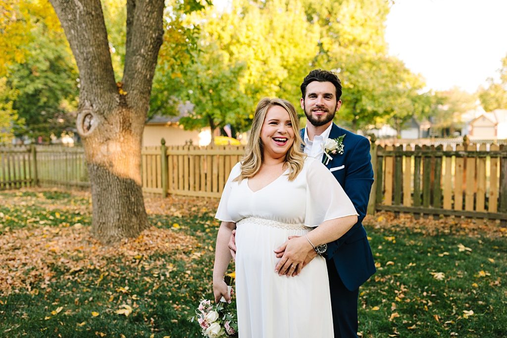 pregnant bride wearing a flowy flutter sleeve wedding dress with a sparkly beaded belt and groom wearing a navy suit in their backyard before their intimate elopement at Raintree Lake in Kansas City