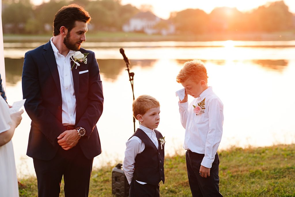 ring bearer cries happy tears during wedding ceremony watching his dad marry his step mom during golden hour at outdoor wedding at raintree lake in lee's summit in kansas city