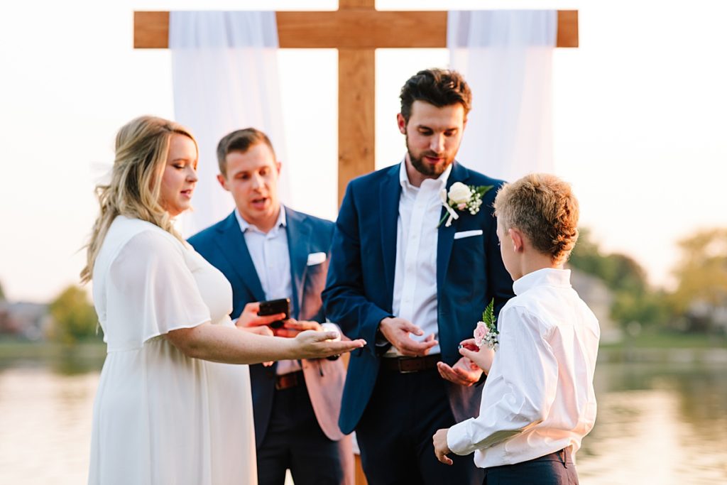 ring bearer hands grooms the rings and wedding bands during outdoor sunset ceremony in october during a warm fall in kansas city