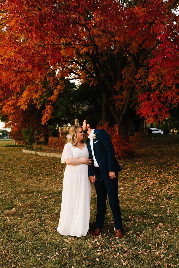 groom kisses bride on the forehead in front of gorgeous tree with red and orange leaves, pose idea for couples photos, From a Large Wedding to an Intimate Elopement at home