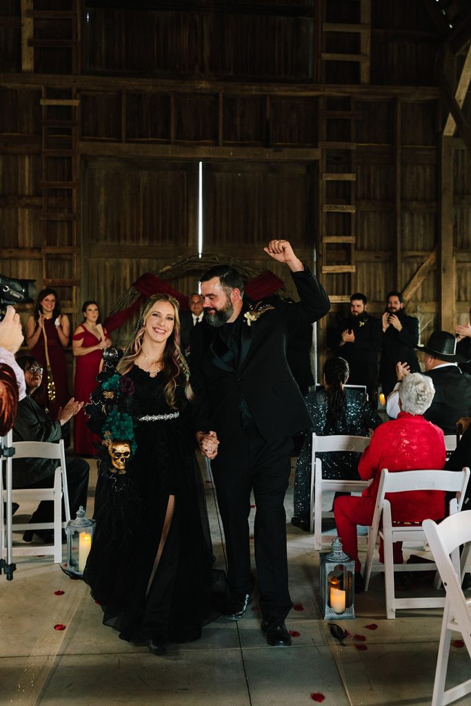 bride and groom celebrate back up the aisle after their halloween wedding ceremony, bride wearing a black wedding dress and holding an alternative bouquet, groom attire is an all black suit