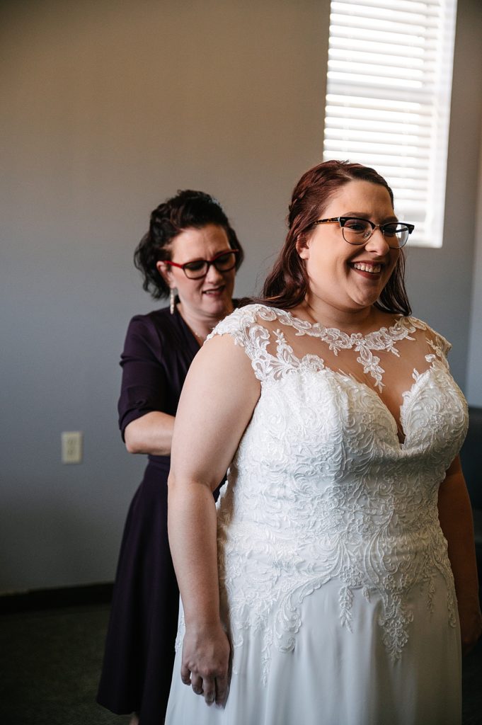 high neck lace wedding dress, bridesmom helps zip bride into dress, bride wearing glasses,