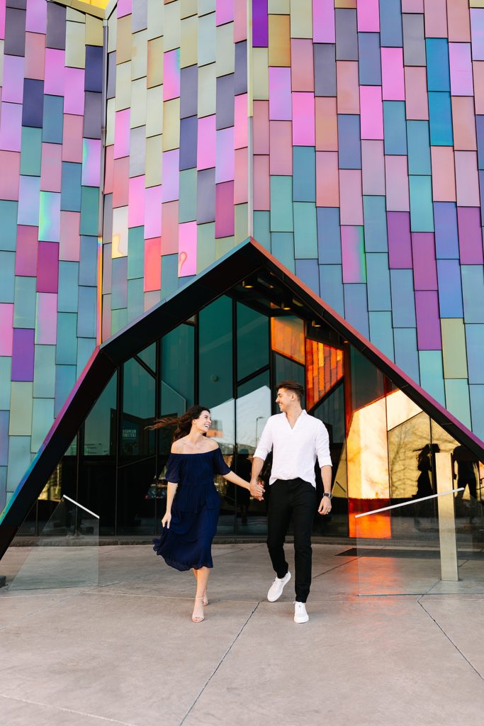Woman in navy dress and man in white shirt walking towards the camera in front of a colorful building. 