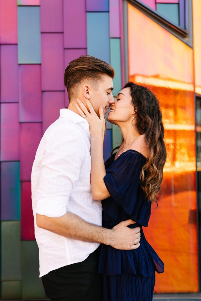 Man and woman about to kiss in front of a colorful building. 