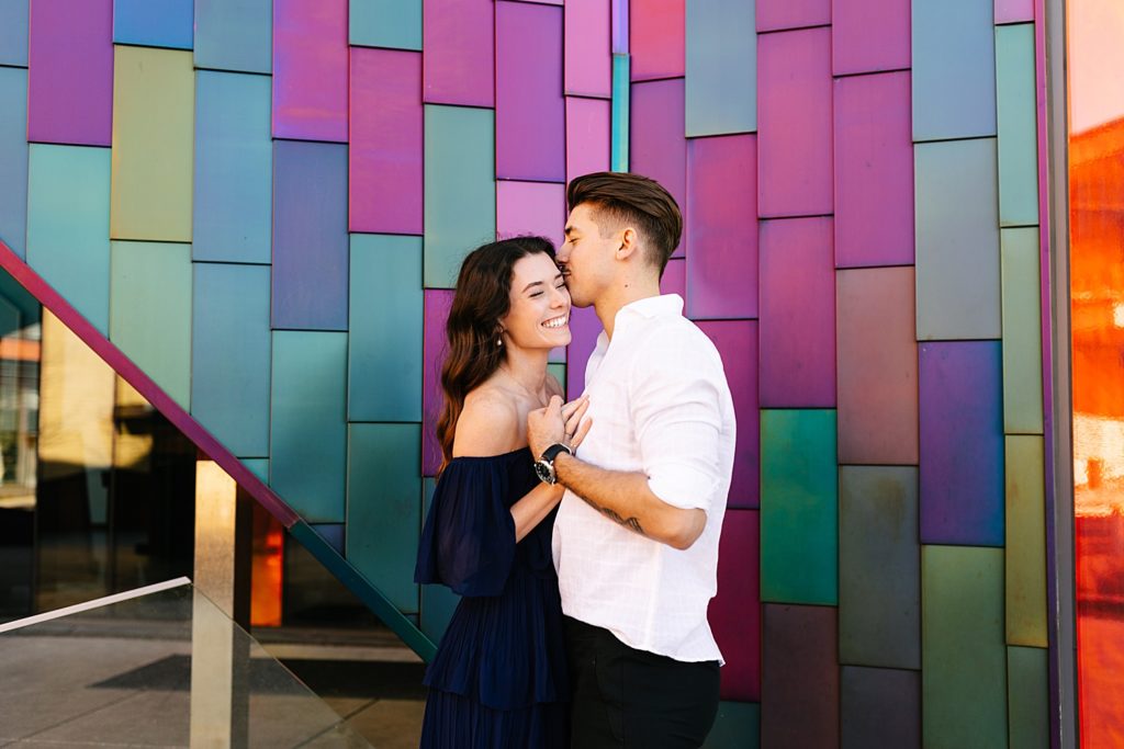Man in white shirt kissing the temple of a brunette woman in a navy dress in front of a colorful wall. 