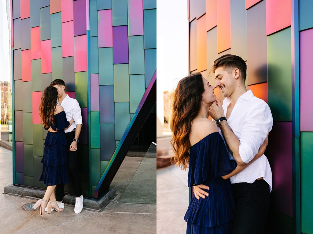 Man leaning against a colorful wall while a woman leans in to kiss him. 