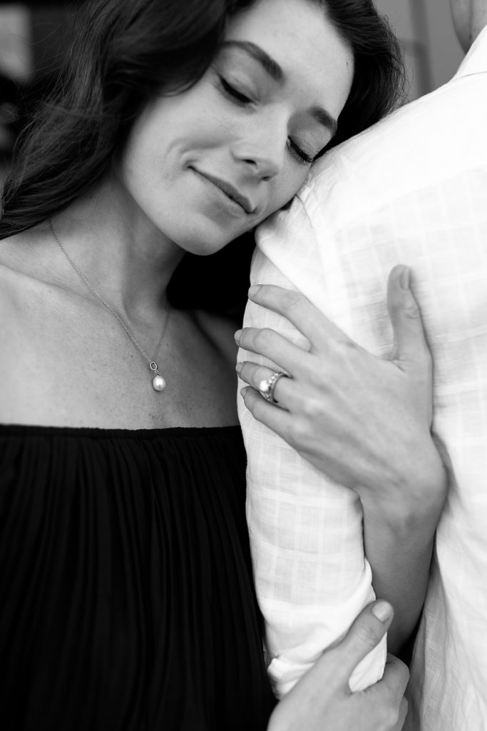 Black and white close up image of a woman snuggling on her fiancé's shoulder with the camera's focus on her pearl necklace. 