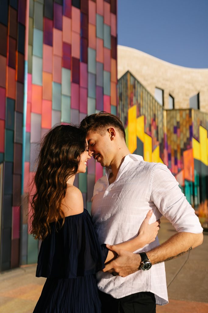 Engaged couple about to kiss in front of a colorful building for their engagement session. 