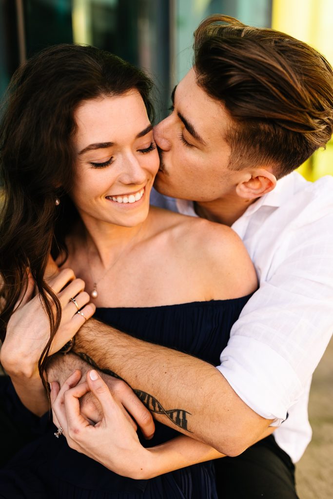 Up close of man wrapping up his fiancé and kissing her check while she is smiling for their engagement session.