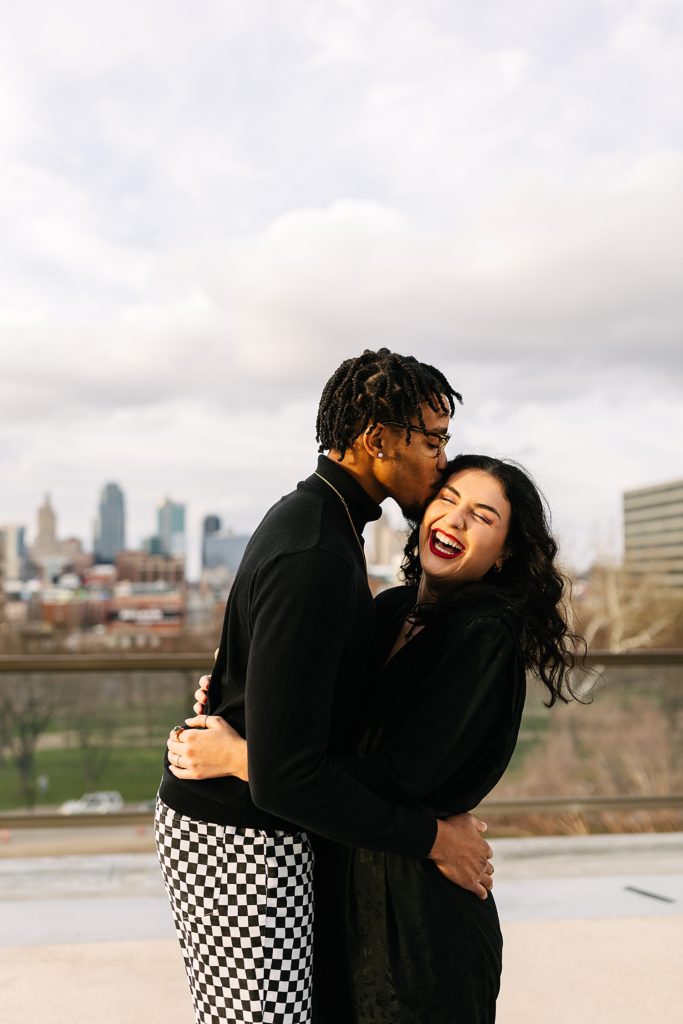 Man kissing woman's temple & embracing her, as she is laughing with the skyline behind them.