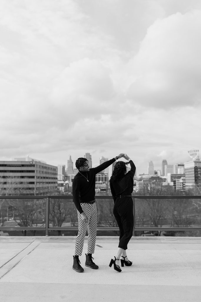 Black and white image of a man and woman dancing on a rooftop with the city skyline behind them. 