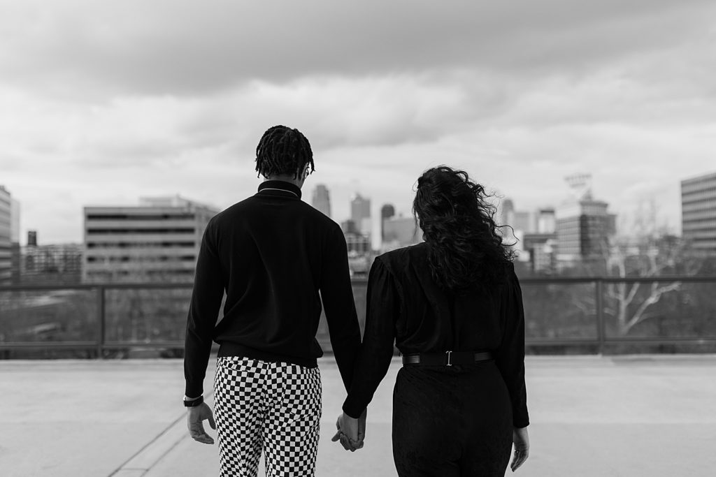 Black and white image of a couple holding hands on a rooftop, facing the city skyline in the distance. 
