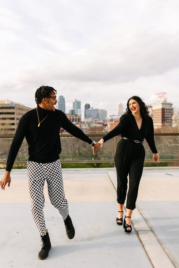 Man and woman laughing and walking towards the camera on a rooftop with the city skyline behind them. 