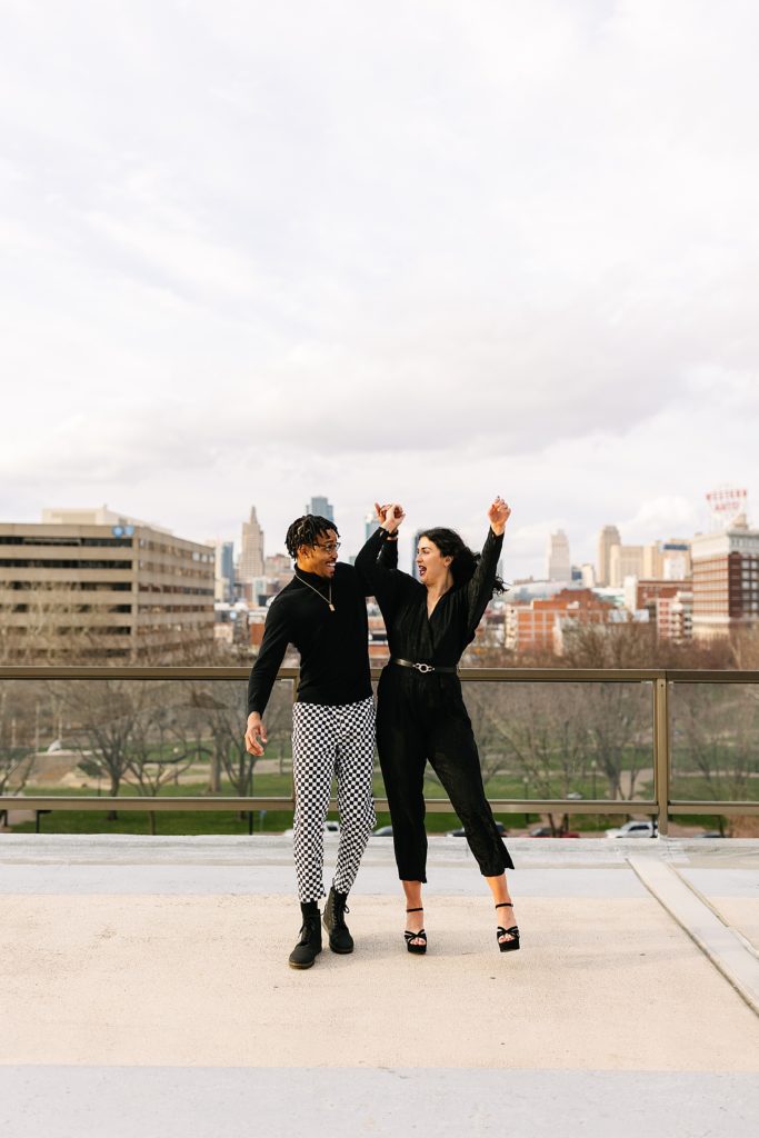 Man and woman bumping hips and laughing with their hands in the air with the city skyline behind them at their rooftop session. 