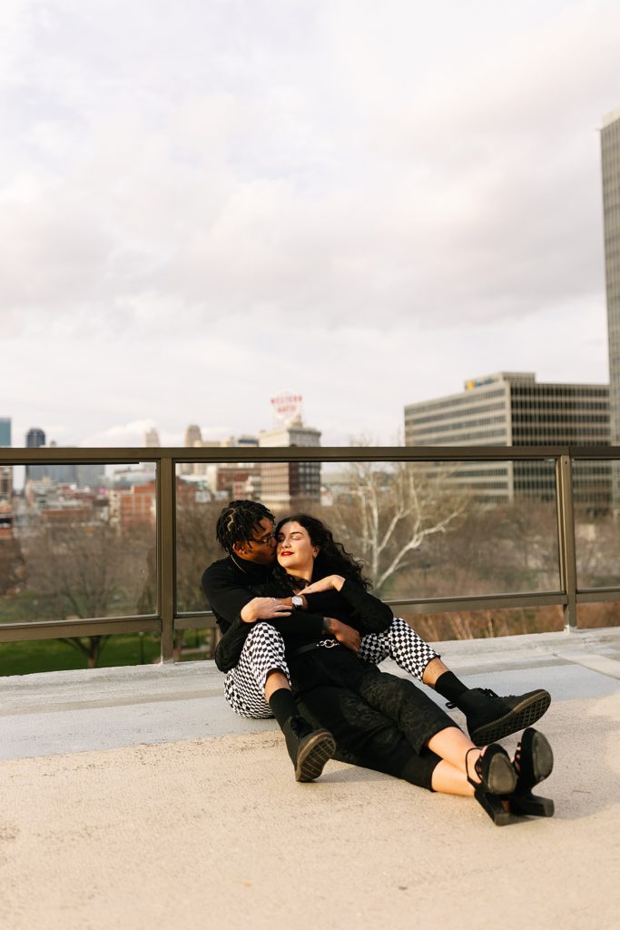 Man and woman curled up sitting on a rooftop. Man is kissing woman's cheek and the background is Kansas City skyline. 