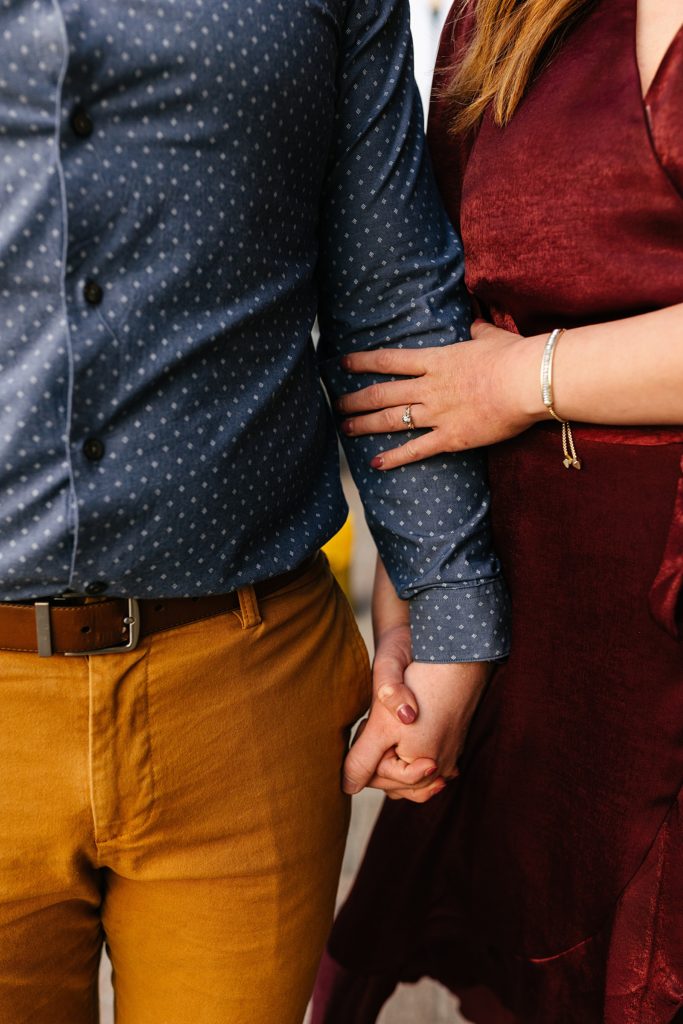 Detail photo of a couple in Fall colors holding hands, the woman has her hand on man's arm with a ring in focus.