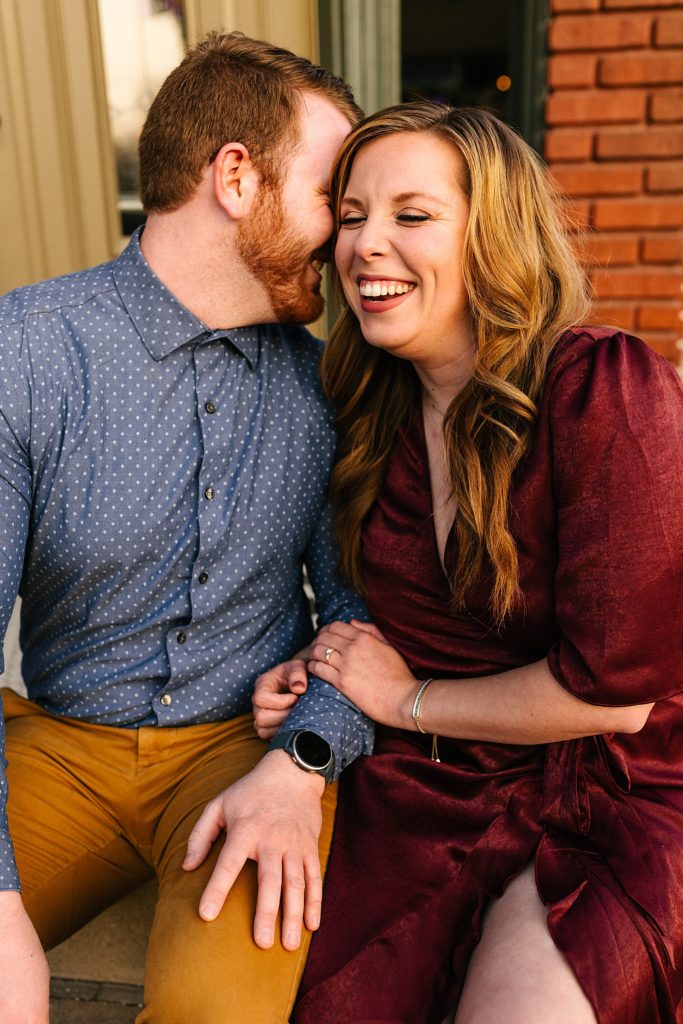 Woman in red dress laughing as her fiancé whispers in her ear.