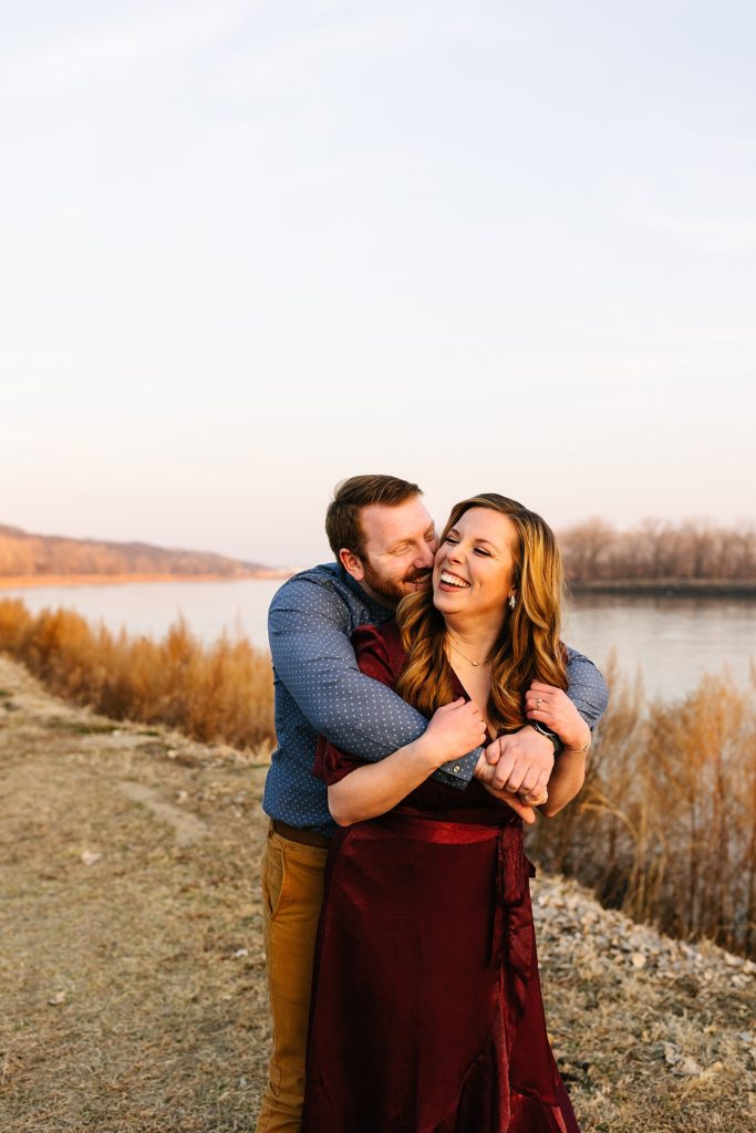 Man wrapping up his fiancé in a hug next to a river in Parkville, Missouri.
