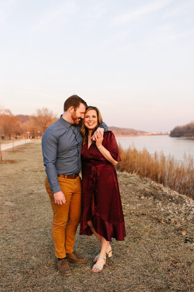 Engaged couple in rich jewel toned clothes embracing next to a river.