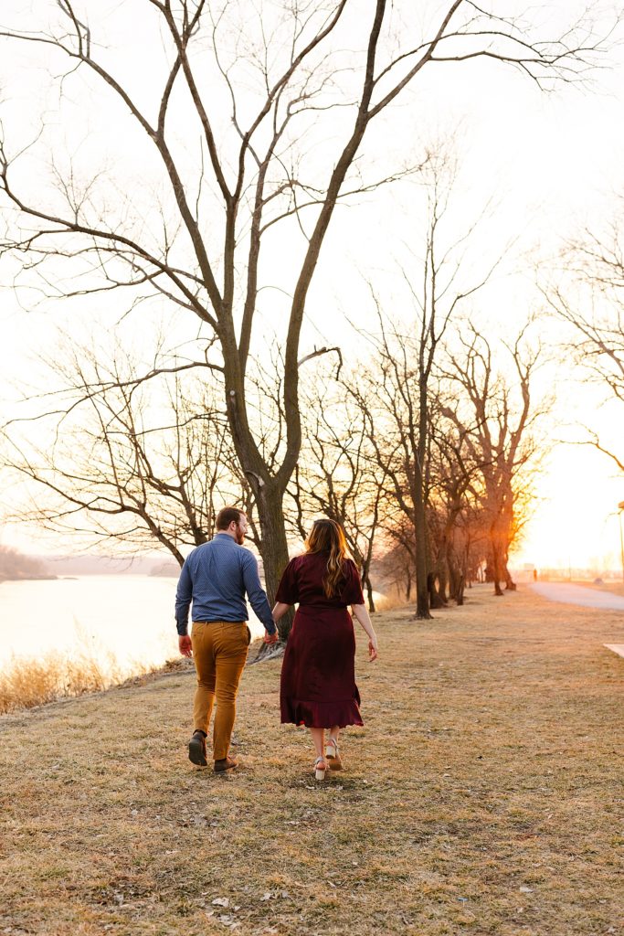 Man in yellow pants and blue shirt, and woman in a red dress, walking away from the camera next to a river in the sunset. 