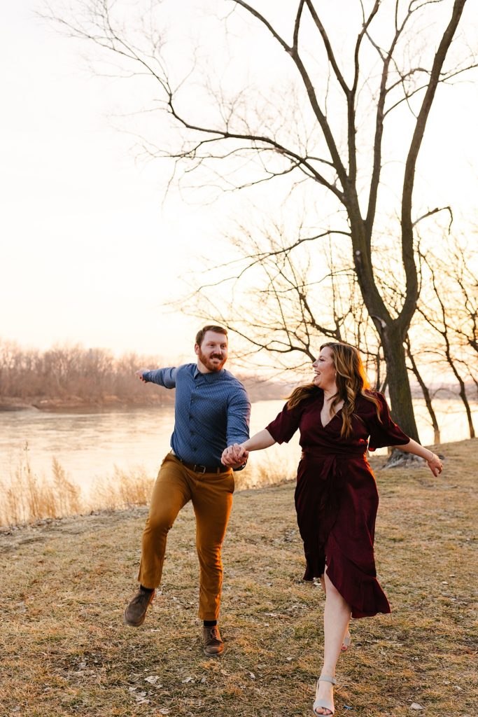 Man and woman in Fall colored clothing dancing and laughing next to a river. 