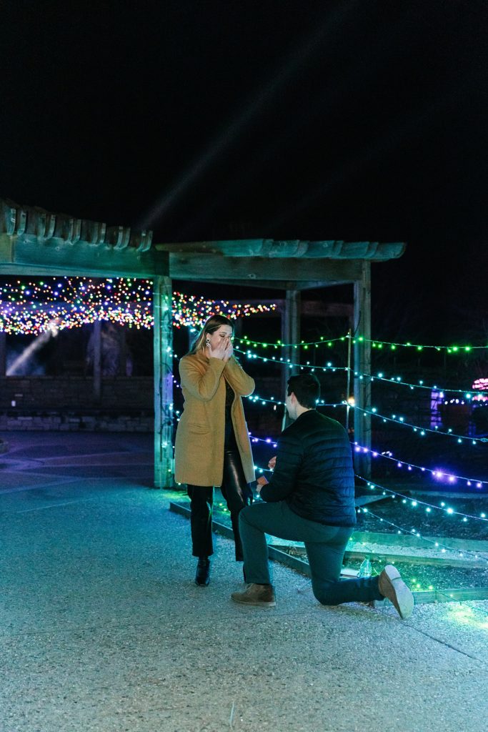 Man down on one knee in front of woman, surrounded by Christmas lights.