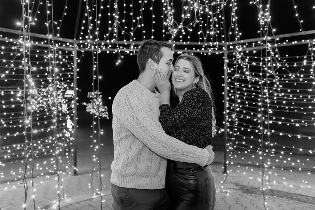 Black and white of man kissing woman's cheek, surrounded by Christmas lights. 