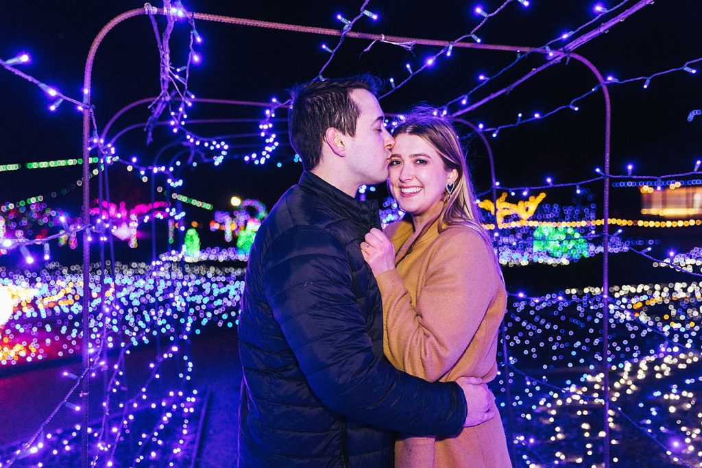 Newly engaged couple embracing under Christmas lights. 
