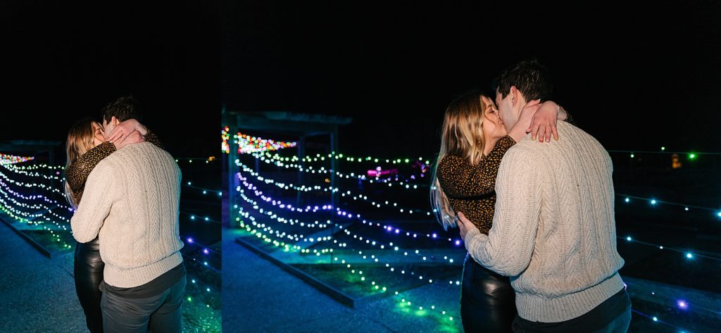 Newly engaged couple kissing in front of Christmas lights.