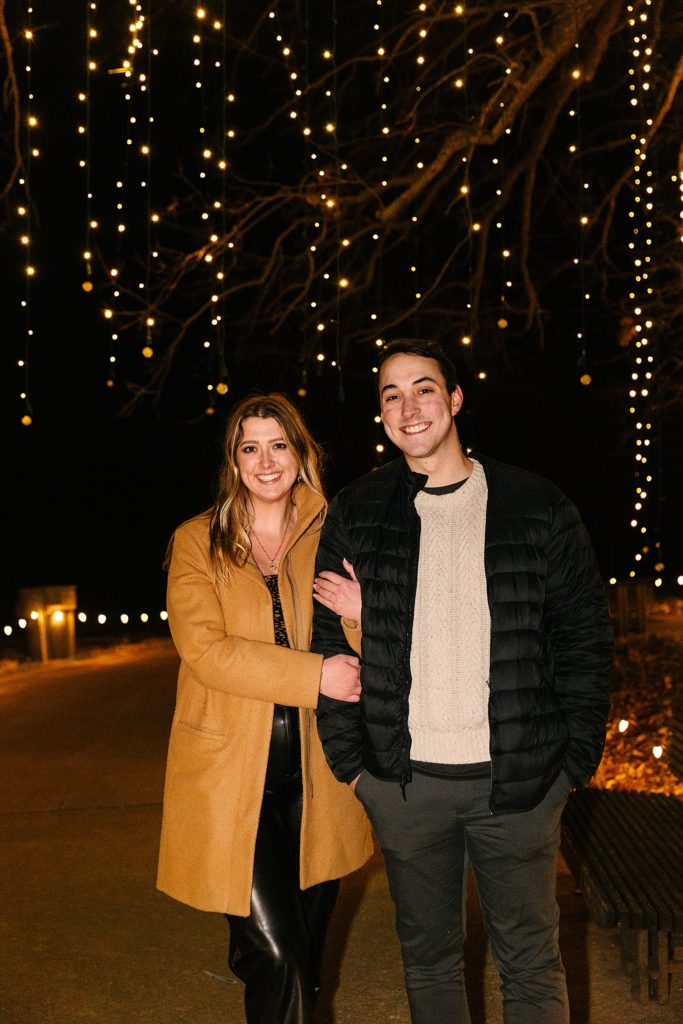 Newly engaged couple posing in front of Christmas lights. 