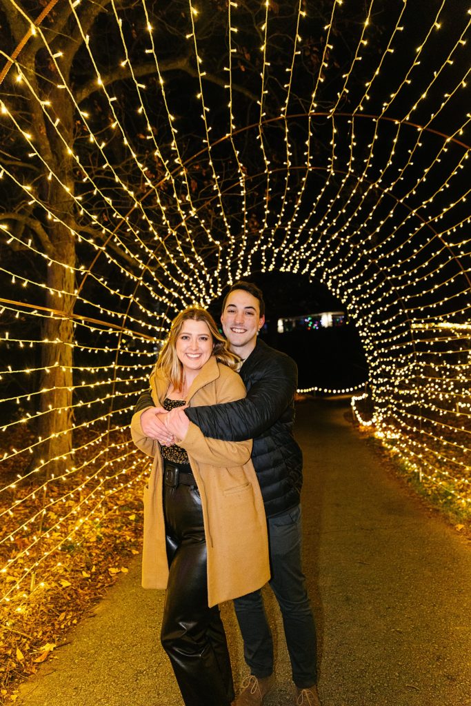 Man wrapping up woman in embrace in a tunnel of Christmas lights. 