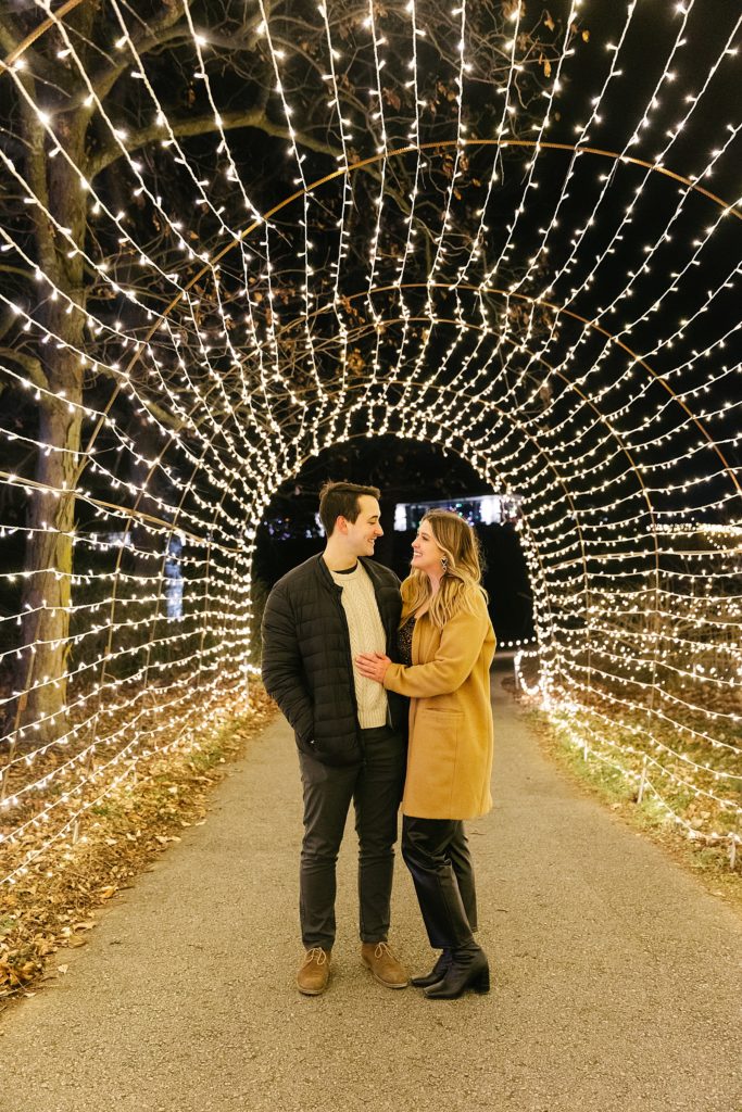 Newly engaged couple looking at each other in a tunnel of Christmas lights. 