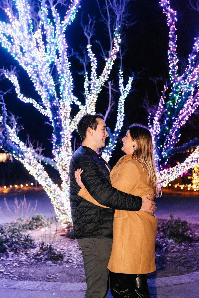 Man and woman embrace in front of Christmas lights. 