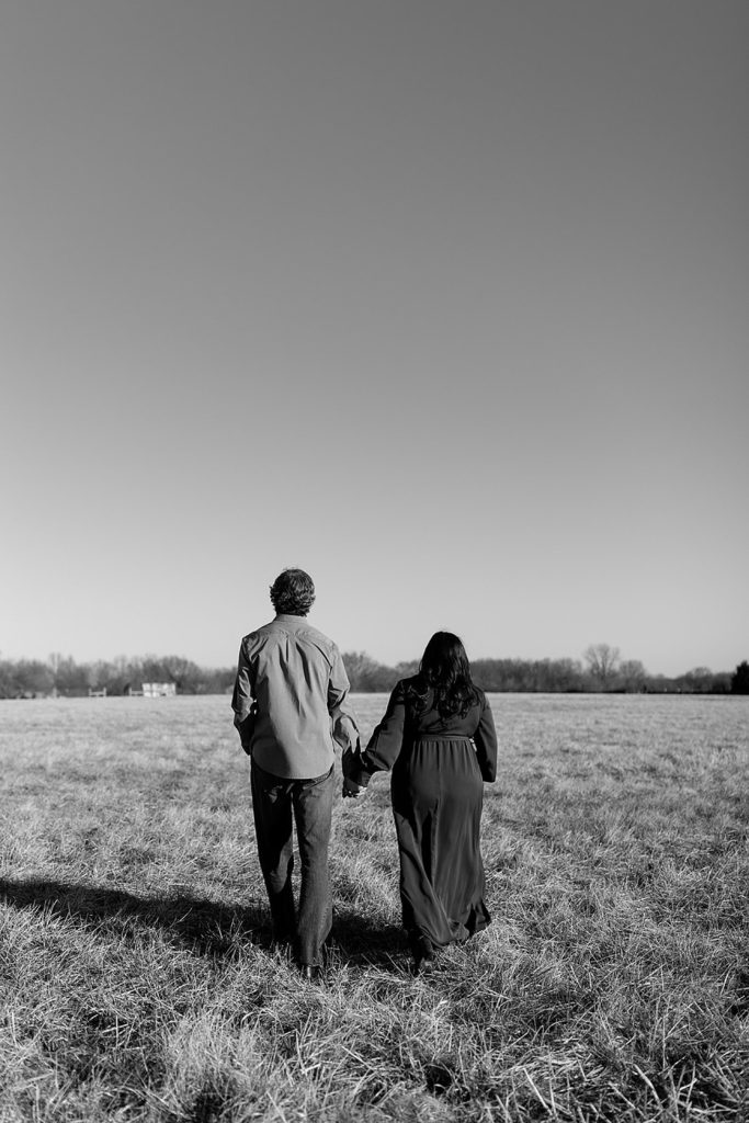 Black and white photo of engaged couple walking away holding hands across a field. 