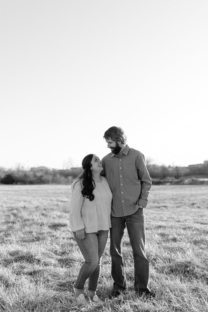 Black and white photo of engaged couple looking at each other in the middle of a field. 