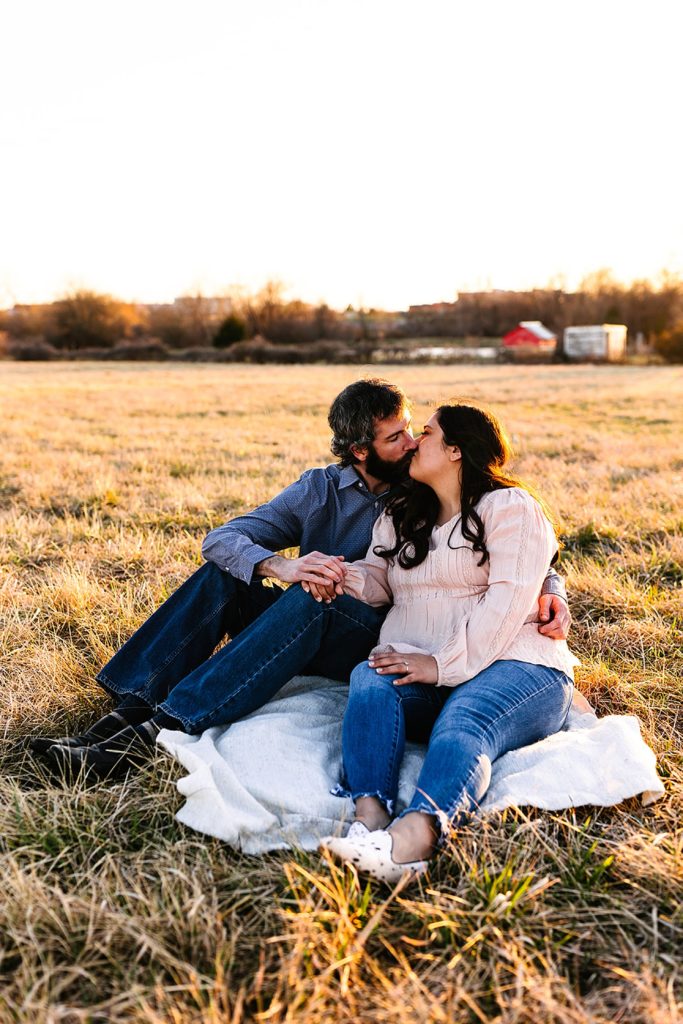 Engaged couple kissing on a picnic blanket in the middle of a field. 