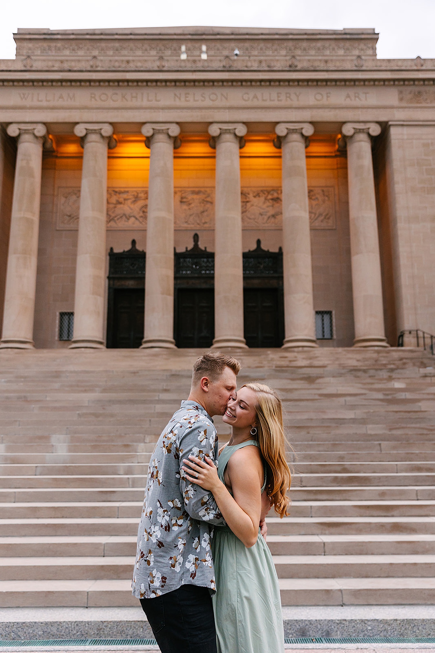 Engaged couple shares kiss in front of the steps at the Nelson Atkins Museum.