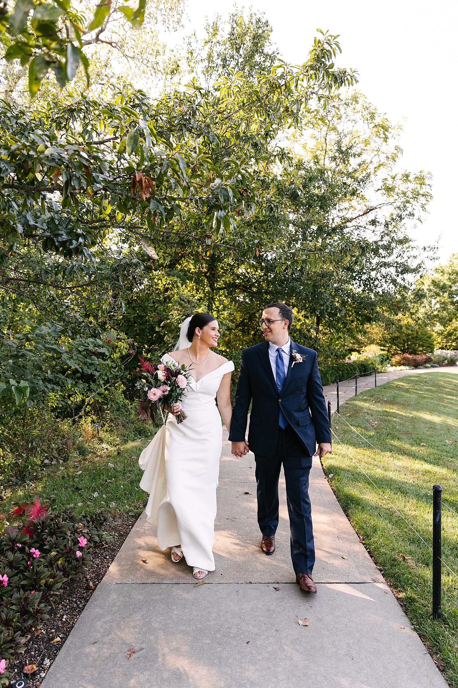 Bride and groom walk pathway at Overland Park Arboretum holding wedding bouquet
